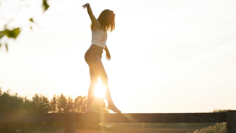 A young woman balancing on a fence during a serene sunrise, exuding freedom and joy.